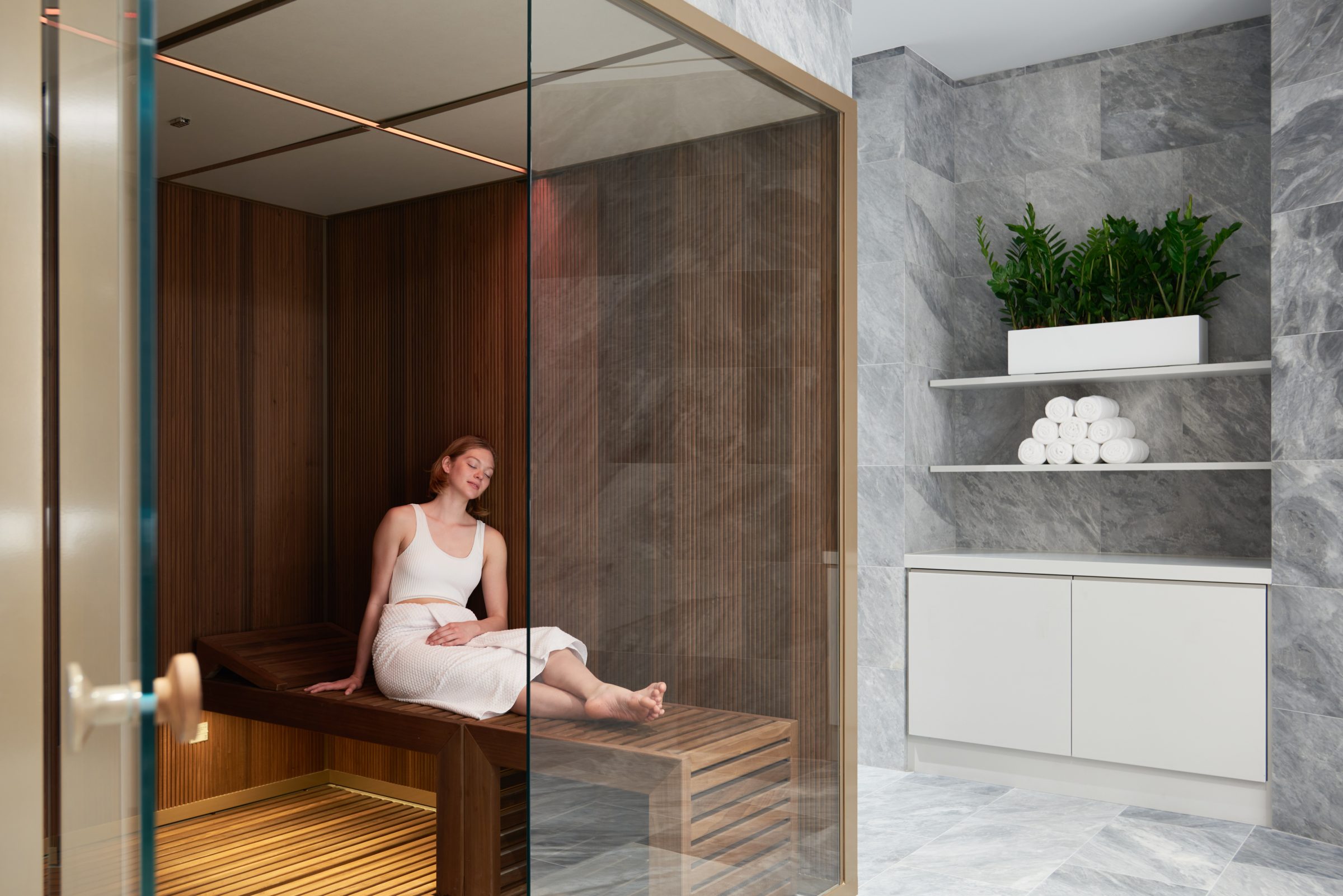A woman relaxing in a glass-fronted sauna beside a marble bathroom.
