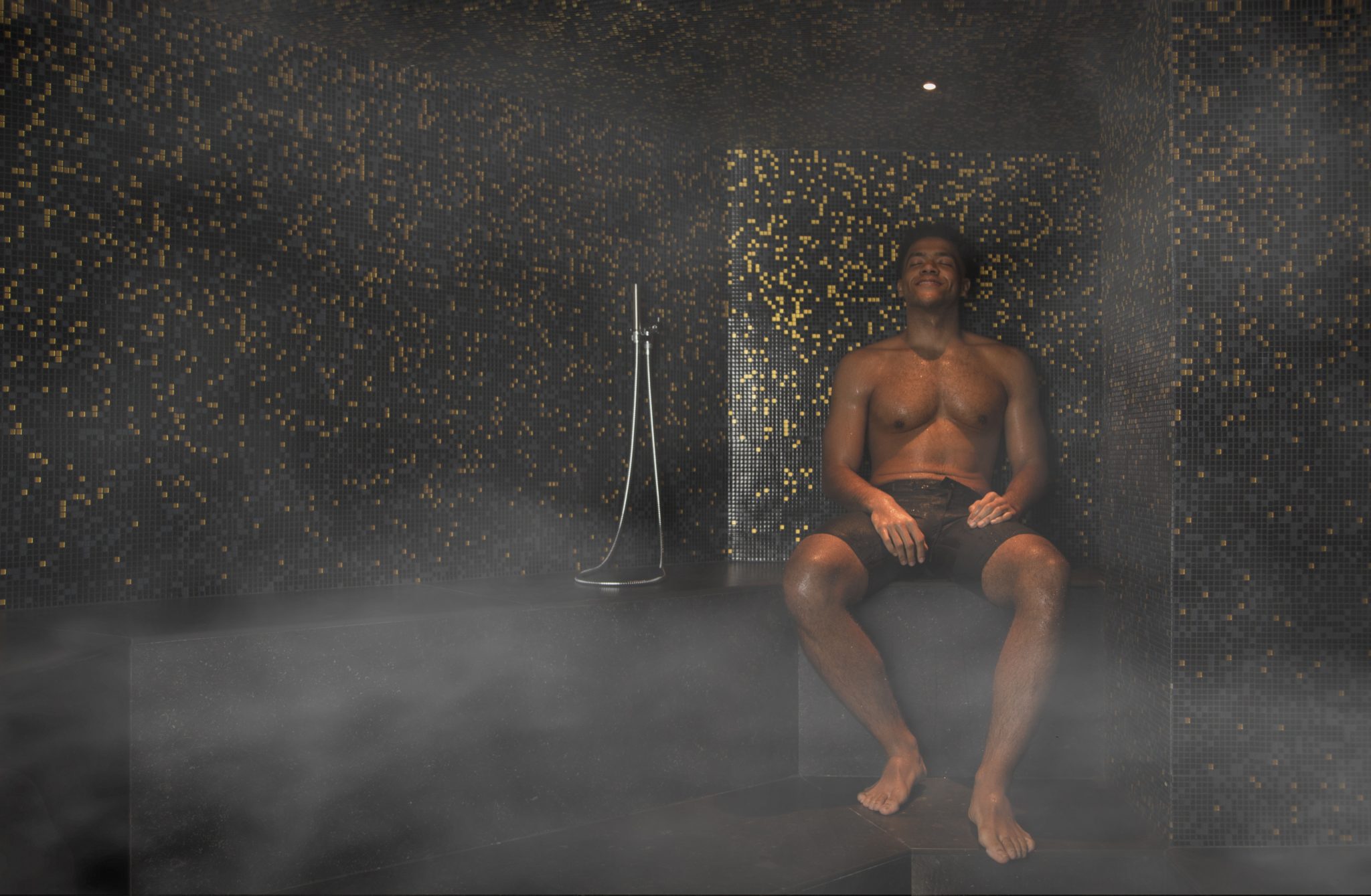 A man seated in a steam-filled room with dark mosaic walls.